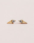 In front of a soft pink background is a pair stud earrings. On one side of each stud is a gold triangle and on the other side is a white diamond. The diamond is on the flat side of the triangle.