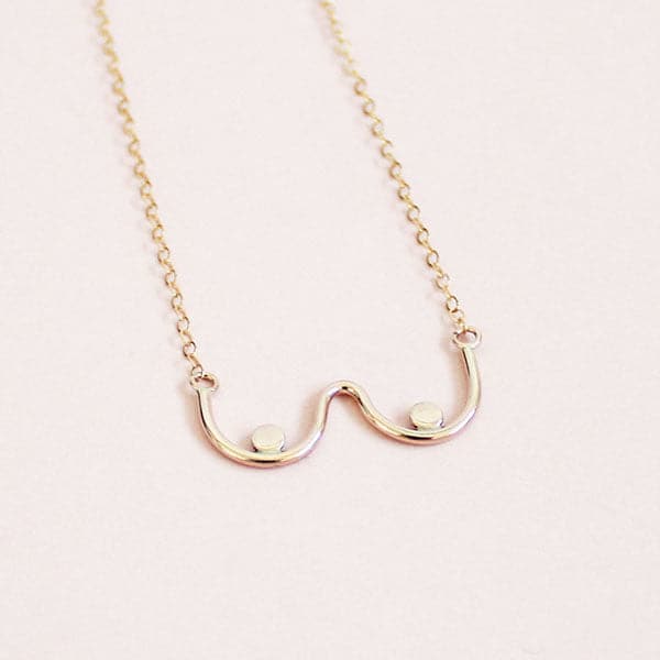 A gold chain necklace with a dainty pendant shaped like the outline of boobs. 