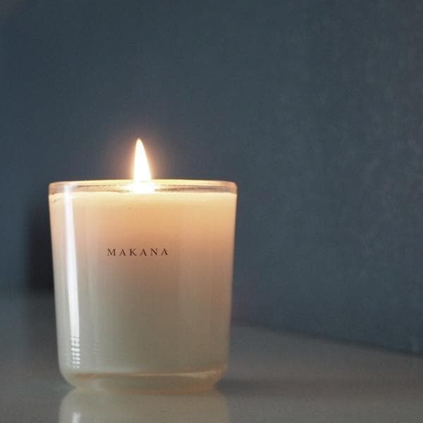 In front of a dark gray background is a clear glass jar. Inside the jar is a white candle with a lit wick in the center. On the front is black text that reads ‘Makana.&#39;