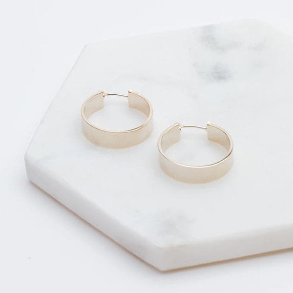 Medium sized silver hoops that have a wider frame and a thin depth. They feature a straight post and a clasp on the back top of the hoop to secure it on your ear.