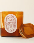 Oval cylinder shaped glass candle with bright tones of burnt orange. The label is an oval shape and reads 'Tobacco & Patchouli' with a galloping horse image within. The middle reads 'PaddyWax Since Ninety Six: Nashville, Tennessee'. Alongside its lid sits propped up against the candle.