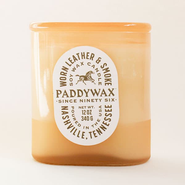 Oval cylinder shaped glass candle with bright tones of soft orange. The label is an oval shape and reads 'Worn Leather & Smoke Soy Wax Candle' in russet capital lettering. The middle reads 'PaddyWax Since Ninety Six' and below 'Nashville, Tennessee'. Within the middle of the label is a print of a galloping horse. 
