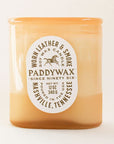 Oval cylinder shaped glass candle with bright tones of soft orange. The label is an oval shape and reads 'Worn Leather & Smoke Soy Wax Candle' in russet capital lettering. The middle reads 'PaddyWax Since Ninety Six' and below 'Nashville, Tennessee'. Within the middle of the label is a print of a galloping horse.