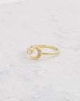 A gold ring with an open front that features a cubic zirconia on one end and a crescent moon on the other, photographed worn on a models hand.