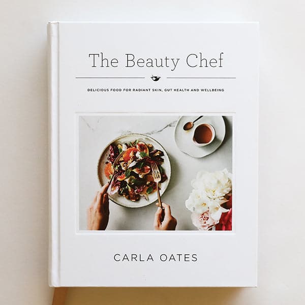 White hardcover of cookbook titled &#39;The Beauty Chef&#39; above an elegant photograph of colorful salad along side white peony flowers and handmade dressing. Below the title reads &#39;Delicious food for radiant skin, gut health and wellbeing&#39; in smaller text. 