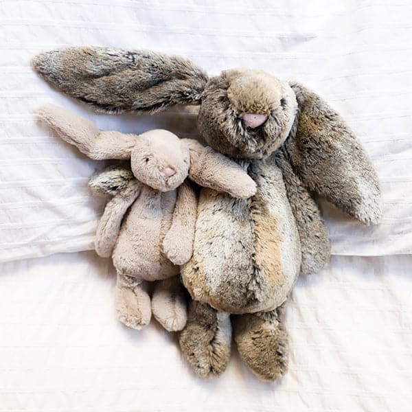 Two, on small and one large, stuffed animal in the shape of rabbits laying on a bed. The small rabbit is beige colored, and the large rabbit is mottled grey and gold.