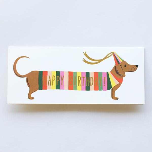A long thin birthday card with a wiener dog illustration on the front with a multicolored stripe sweater on with gold lettering across it that reads, "Happy Birthday". The dog is also wearing a multicolored birthday hat with two ribbons hanging from it. The card comes with a yellow envelope photographed here. 
