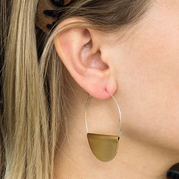 Photo of the side of a woman&#39;s face showing her ear. She is wearing a pair of Hathor hoop earrings. The earrings have a thin silver wire hoop with a half circle of brass folded over the bottom of the wire circle.