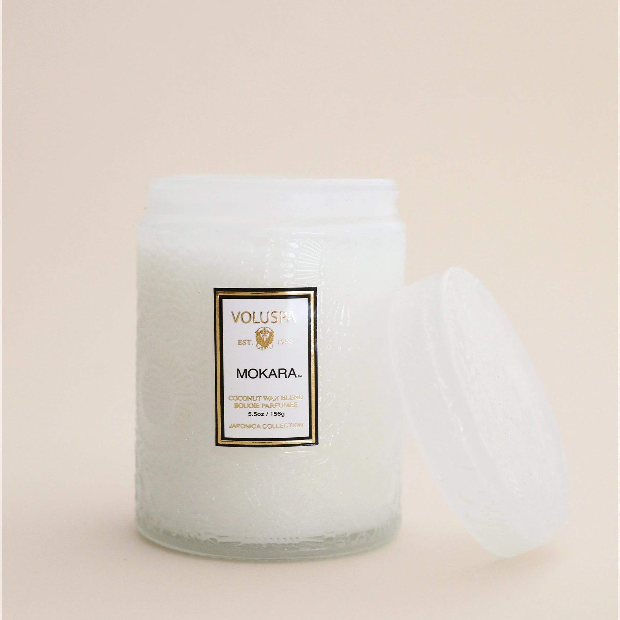 A decorative glass jar candle in a white color with a rectangular label on the front that reads, "Voluspa Mokara" with a coordinating decorative glass lid.