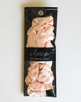 In front of a white background is a black rectangular package. It has a clear top with satin, blush pink scrunchies inside. Around the middle is a horizontal black band with white text that reads ‘sleep, satin sleep scrunchies.’