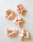 On top of a white background is five blush pink, satin scrunchies.