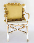 Against a white background is a white and brown wicker chair. On the chair is a mustard colored square pillow. The pillow is lined with small balls with tassels.