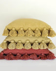 Against a white background is a stack of three square pillows. The edge of all of the pillows are lined with small balls with tassels. The top two pillows are mustard and the bottom pillow is rust.
