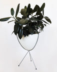 In front of white background is a white planter in a silver metal stand. The bullet planter is wide at the top and narrow at the bottom. The metal stand has three legs. Inside the planter is a deep green rubber tree.