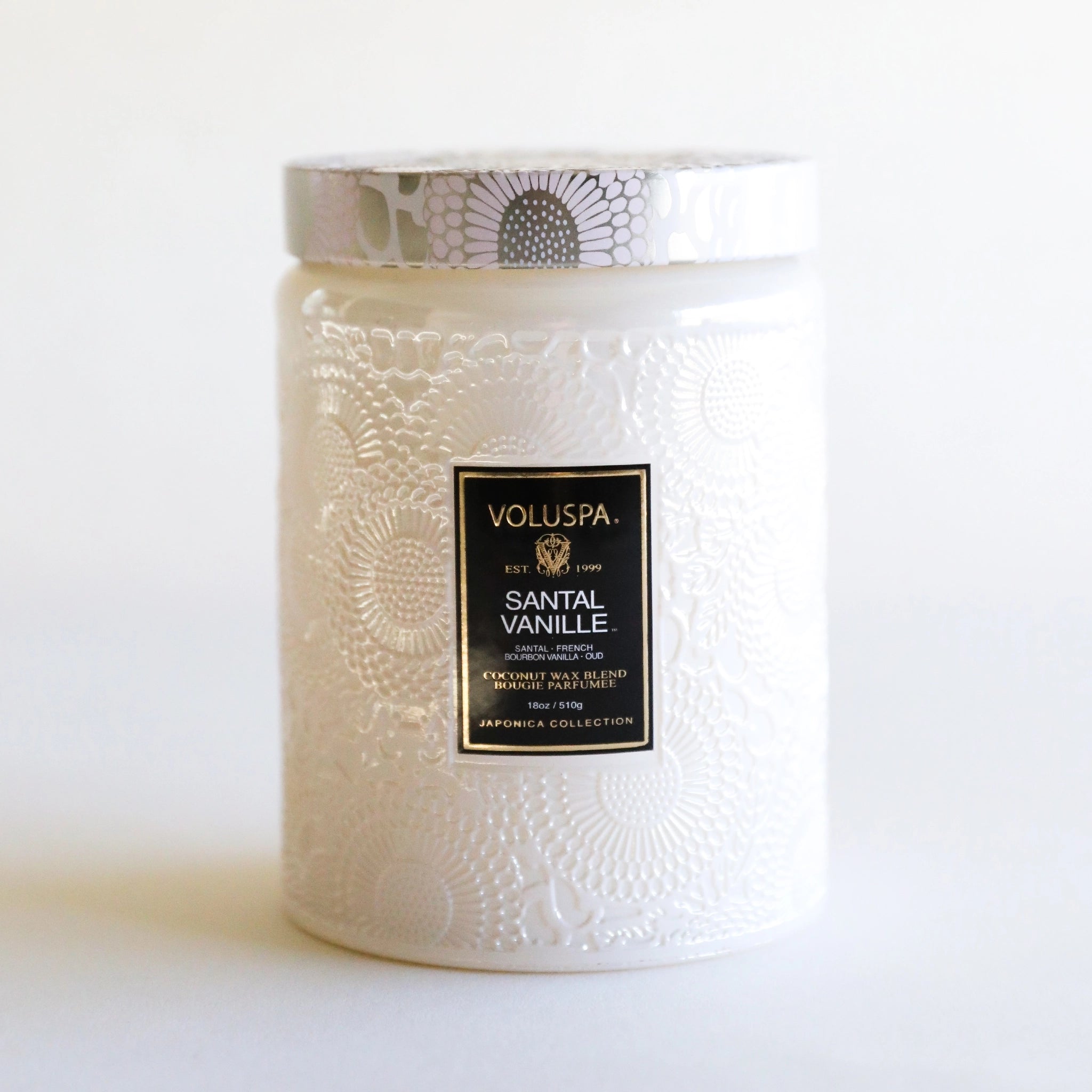 In front of a white background is a large light pink glass jar. The glass jar has a floral pattern all over it. In the middle is a black rectangular sticker with gold text at the top that reads ‘voluspa.’ Below is white text that reads ‘santal vanille.’ On top of the candle is a roun tin lid. The lid has a light pink, silver and lavender flower pattern on it.