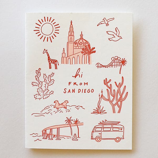 Photo of a greeting card that reads &quot;hi from san diego&quot; in rust letters on a white background. There are also rust colored simple line drawings of San Diego landmarks surrounding the writing, including a sun, the Coronado bridge, La Jolla beach seals, cacti, Balboa Park, and a giraffe representing the zoo.
