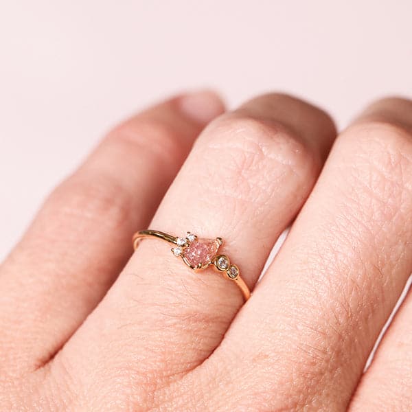Against a soft pink background is a person’s hand with the ring on their left ring finger.  On the front of the ring is a light pink crystal tear drop. Lining the left side of the tear drop are three white, round crystals. To the right of the tear drop is two white crystals with a gold border.