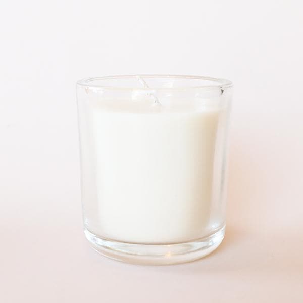 In front fo a white background is a round glass jar. Inside the jar is a white candle with a white wick in the center. 