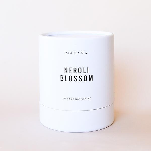On a cream background is a white cardboard candle container with a clear glass jar candle inside with a single wick candle inside along with black text on the front that reads, &quot;Neroli Blossom&quot;.