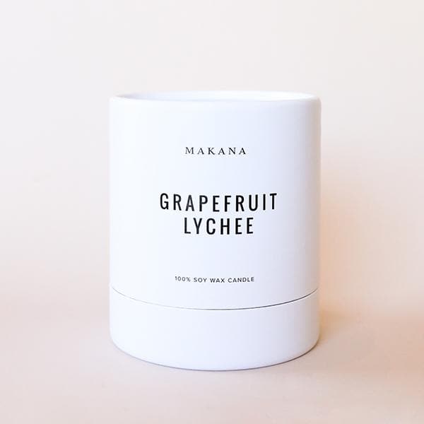 On a cream background is a white candle with black text that reads, &quot;Makana Grapefruit Lychee 100% Soy Wax Candle&quot;. 