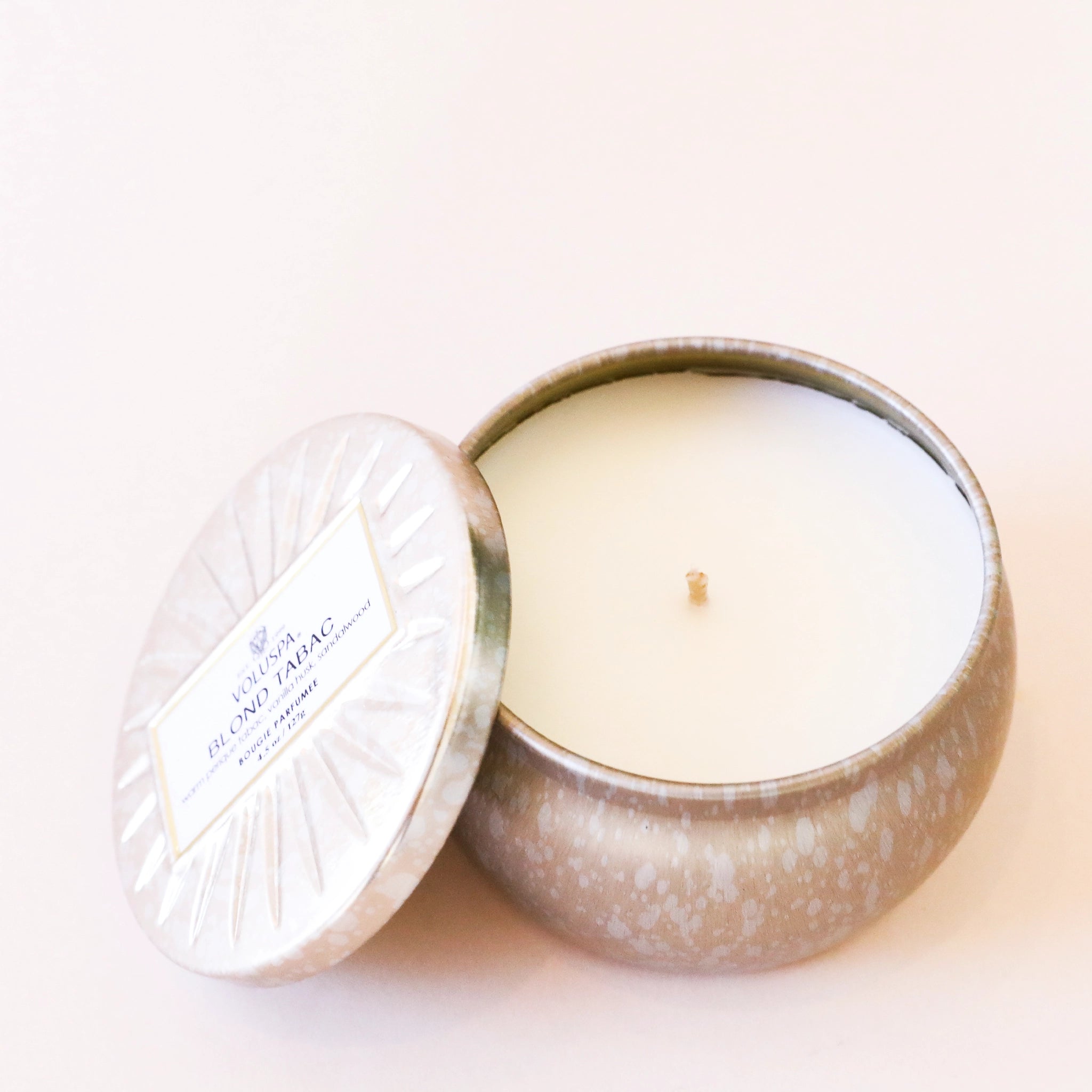 On a cream background is a light silver and gold mini tin candle with white wax and a single wick. Also included is the matching lid. 