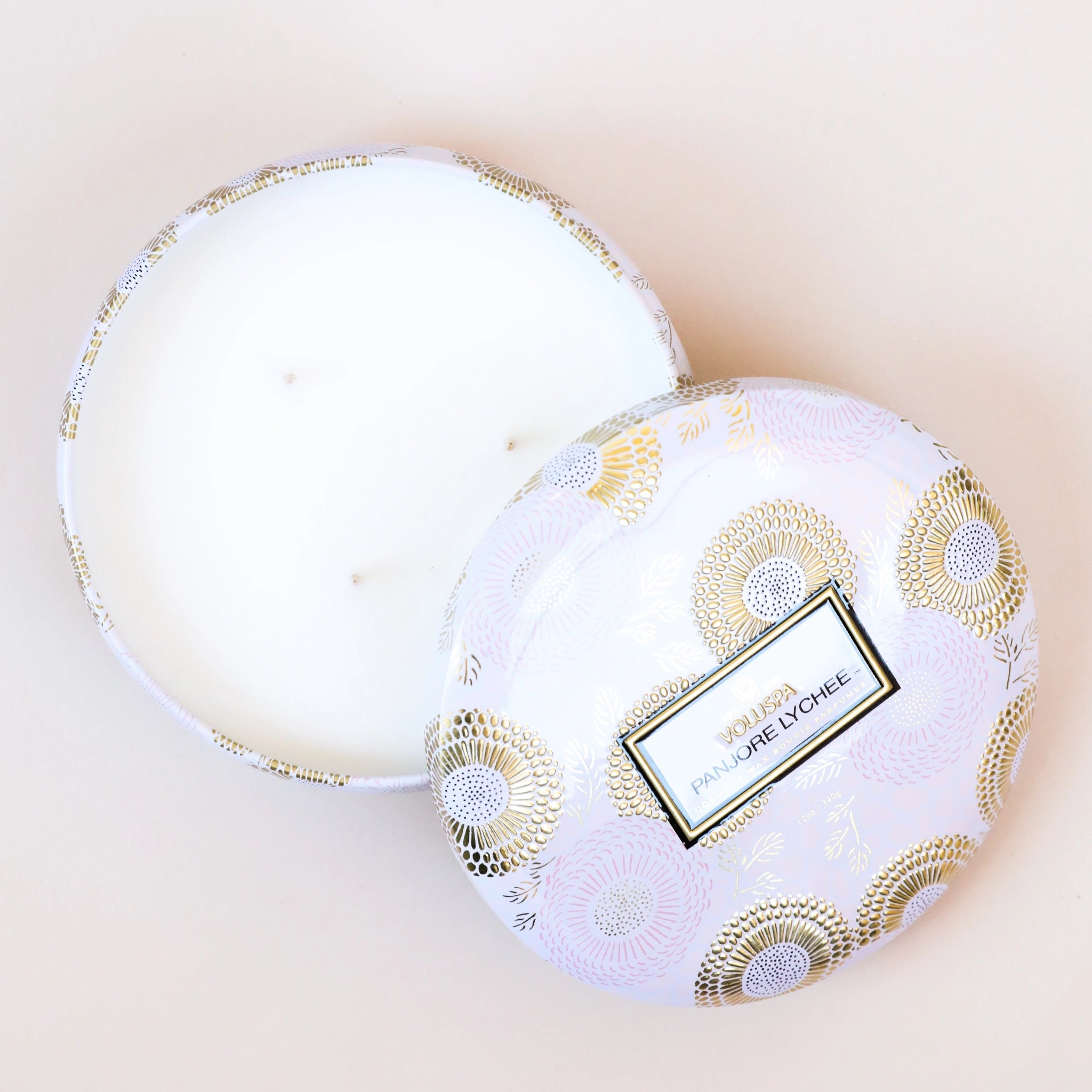 Birds eye view of a short, round tin. Inside the tin is a white candle with three white wicks. Leaning against the right side is a round tin lid. The lid has a light pink and gold floral pattern on it. In the middle is a white sticker with gold and black text inside.