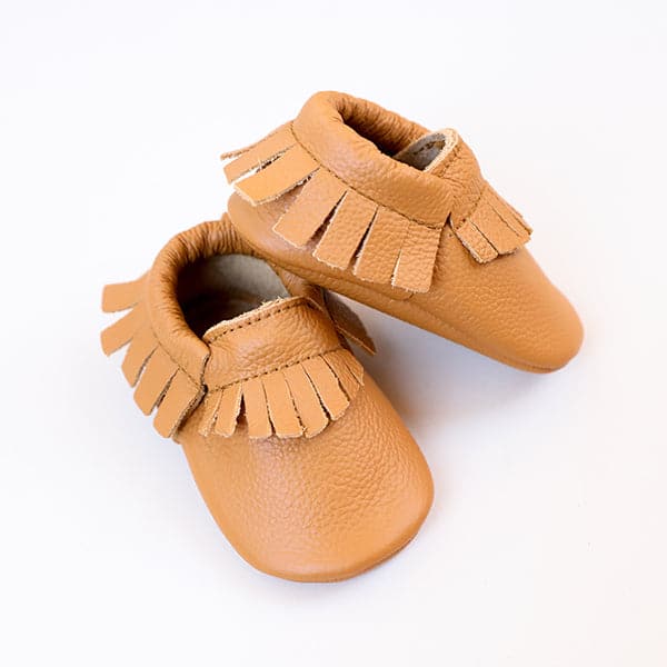 On a white background is a pair of tan baby moccasins that have a fringe detail around the edges. 