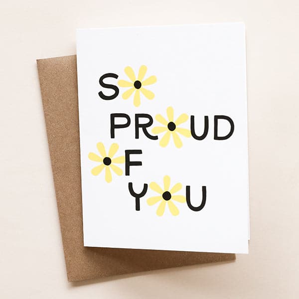 On top of a brown envelope is a white card. In black text it reads ‘so proud of you.’ The ‘o’ in each word is a yellow daisy with a black center. 