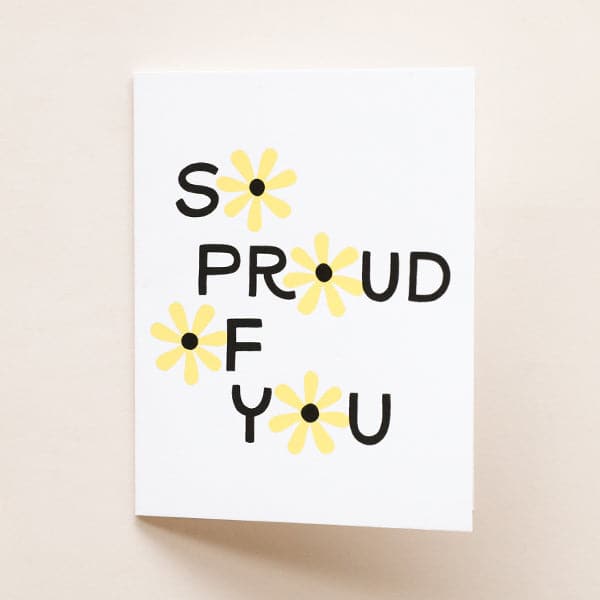 In front of a tan background is a white card. In black text it reads ‘so proud of you.’ The ‘o’ in each word is a yellow daisy with a black center. 