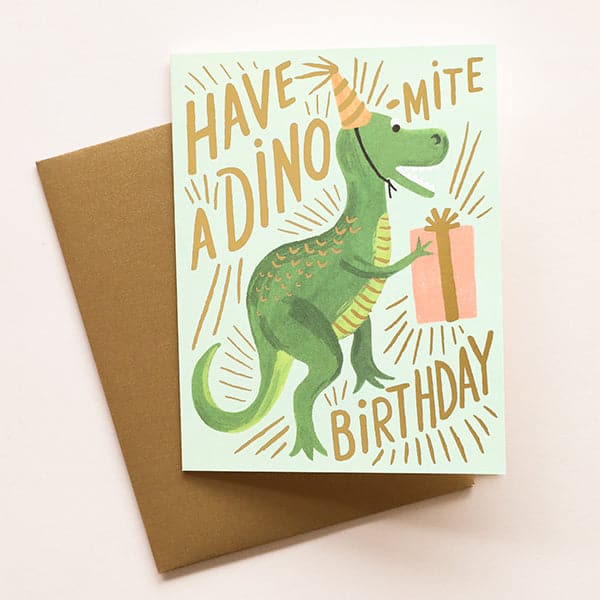 A mint card with gold letters that read, &quot;Have A Dino-Mite Birthday&quot; with a illustration of a green t-rex dinosaur wearing a yellow striped birthday hat and holding a pink birthday gift as well as a coordinating envelope.
