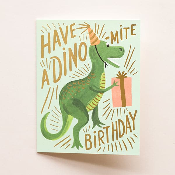 A mint card with gold letters that read, &quot;Have A Dino-Mite Birthday&quot; with a illustration of a green t-rex dinosaur wearing a yellow striped birthday hat and holding a pink birthday gift as well as a coordinating envelope.
