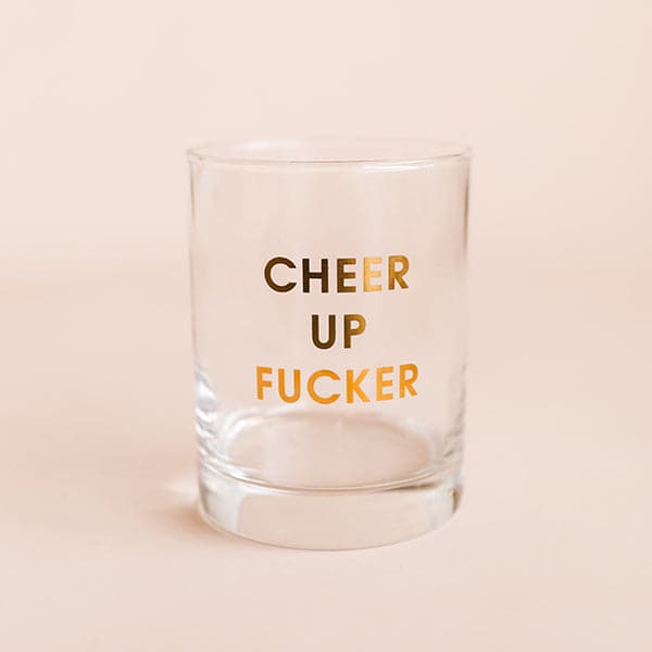 A glass tumbler with gold foil lettering that reads, "Cheer Up Fucker".