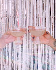 Two iridescent coup glasses with ribbed bases held in front of a tinsel background.