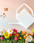 An ivory plastic watering can with a narrow spout and square handle and a rectangle breeze block design on the sides. This photograph has the watering can staged above a bed of pink, yellow and white florals and two butterflies on the left hand side.