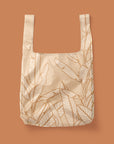 Photographed in front of a terracotta background is a light tan / pink nylon reusable bag with a cream outlined Banana Leaves print on the front and back.