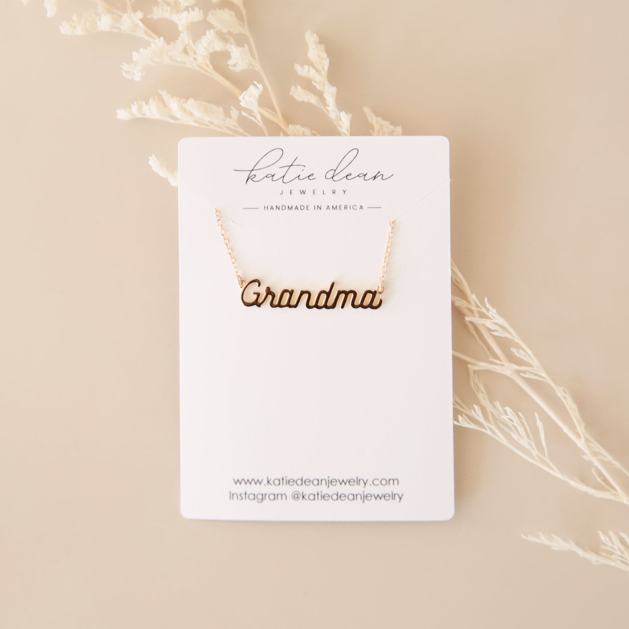 A dainty gold chain necklace with gold letters that read, "Grandma" as a pendant on a white cardboard rectangle that reads, "Katie Dean Jewelry".