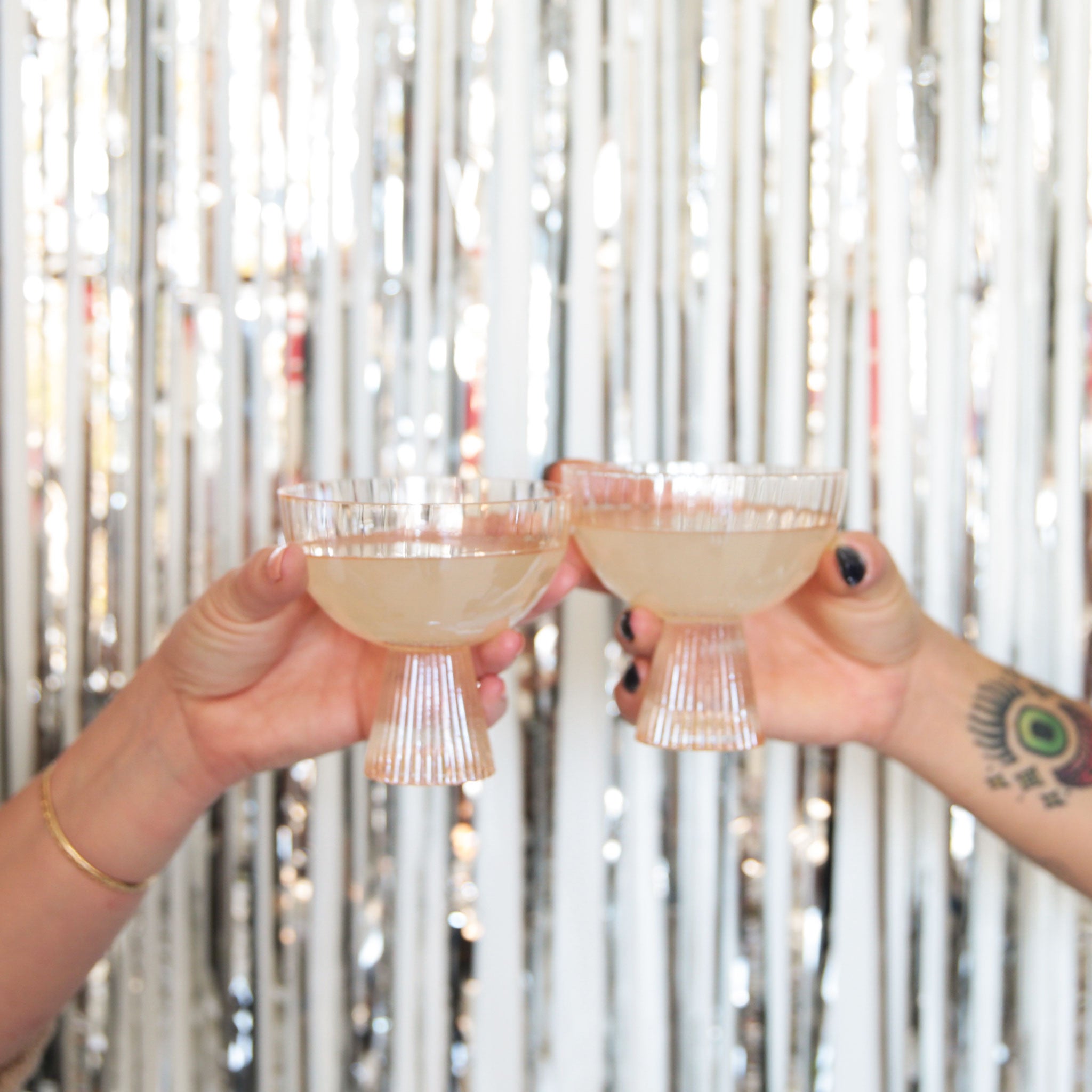 Two light peach beveled coupe glasses with a ribbed stem and a wider ribbed cup being held up doing a cheers in front of a silver tinsel background.