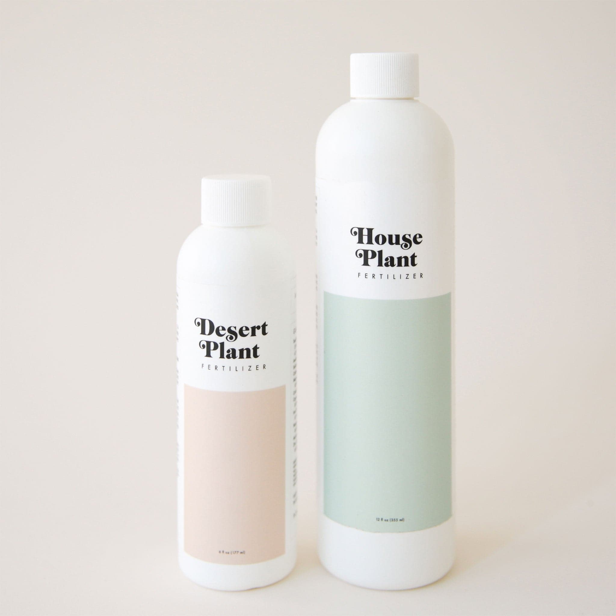 On a cream background is a white plastic bottle of fertilizer with a light teal rectangular shape on the bottom half and text on the top that reads, &quot;House Plant Fertilizer&quot; in black letters photographed here next to a smaller bottle of Desert Plant Fertilizer. 