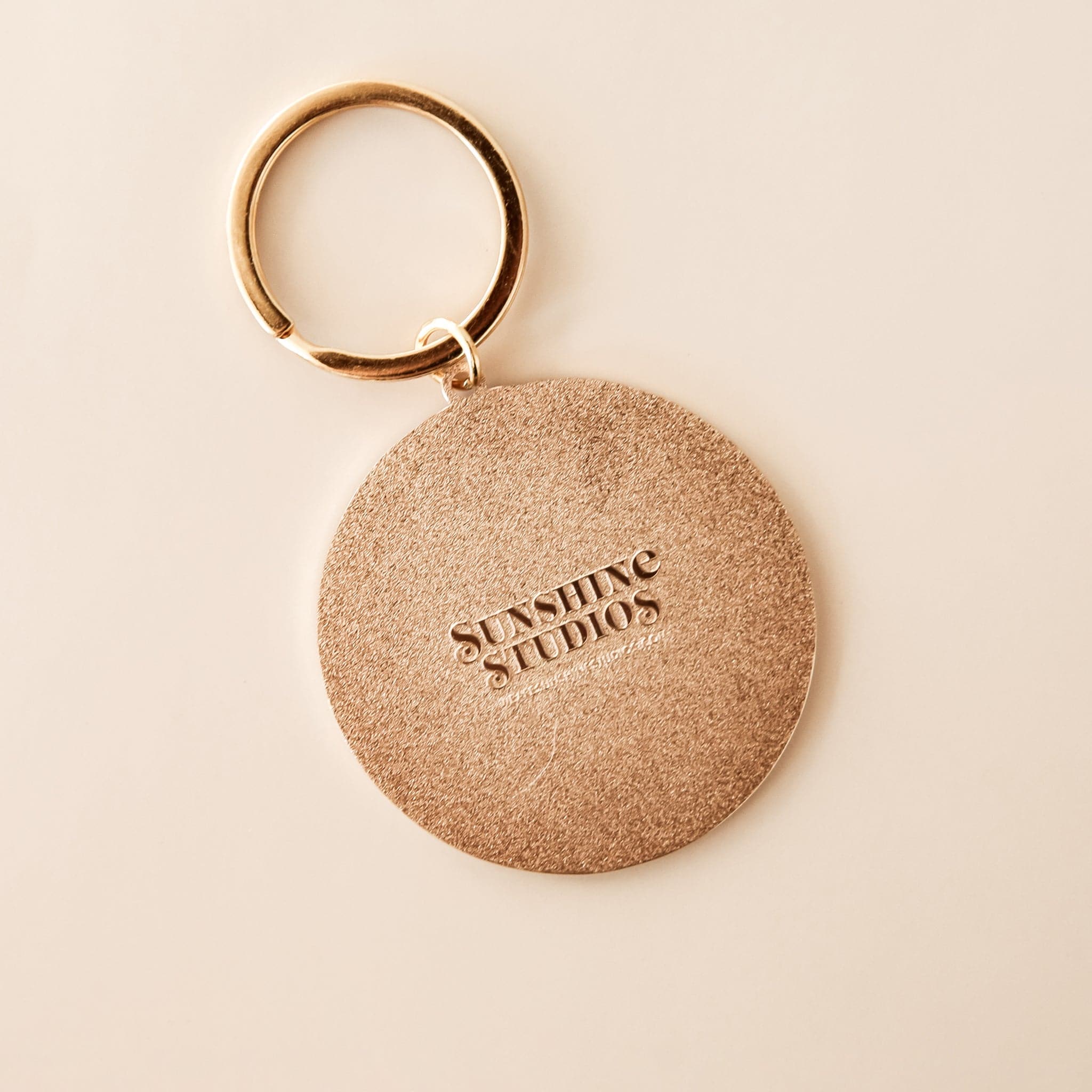 Textured backside of round keychain reading &#39;sunshine studios&#39; in raised lettering. The keychain is complete with a golden keychain loop.