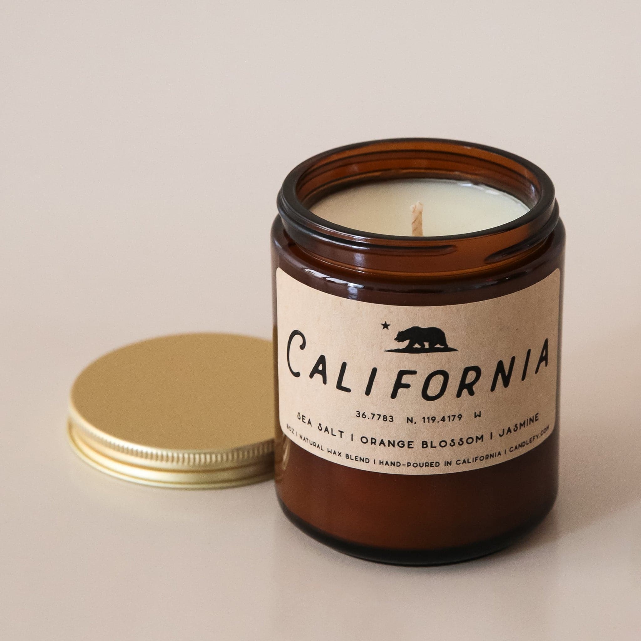 An amber glass jar candle with a light tan rectangular label that reads, &quot;California Sea Salt | Orange Blossom | Jasmine |&quot; in black letters along with a black filled in image of the California state bear and a gold screw on lid.