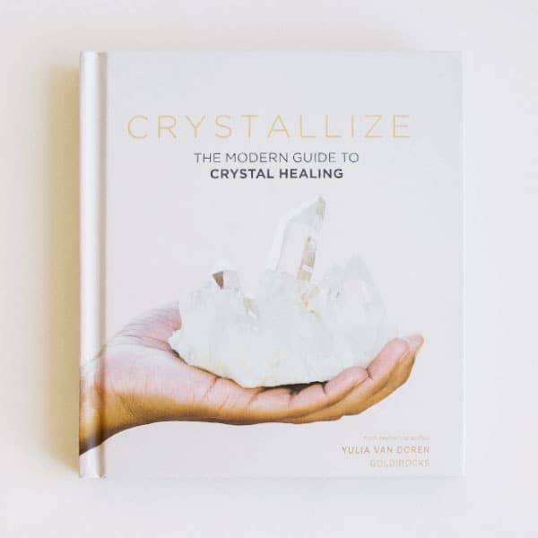 On an ivory background is a light pink book cover with a photograph of a model holding a large clear quartz crystal and the title above it that reads, "Crystallize The Modern Guide To Crystal Healing".
