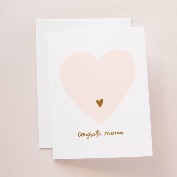 On a light pink background is a white card and envelope with a light pink heart in the center and a smaller gold heart inside of that along with gold text that reads, "Congrats Mama" along the bottom.
