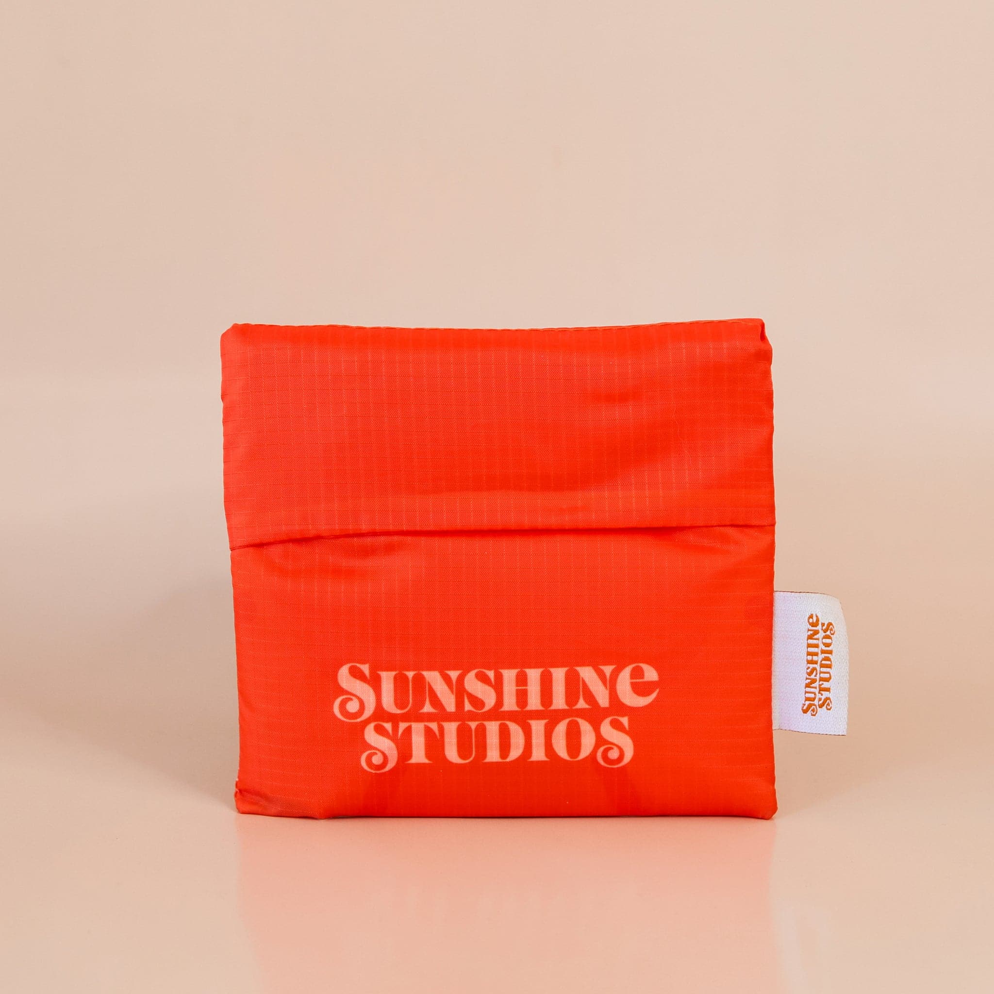 Red-orange nylon bag folded up into a compact square. The bag reads &#39;sunshine studios&#39; in off white lettering and has a small white tag that reads &#39;sunshine studios&#39; in orange lettering.
