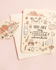 A cream colored card with a bunch of desert illustrations, including Joshua trees, cacti, stars, a camping trailer and more along with the words, "You Are My Forever Adventure". Also included is a white envelope and a small sticker with a camping chair and trailer against a desert mountain range.