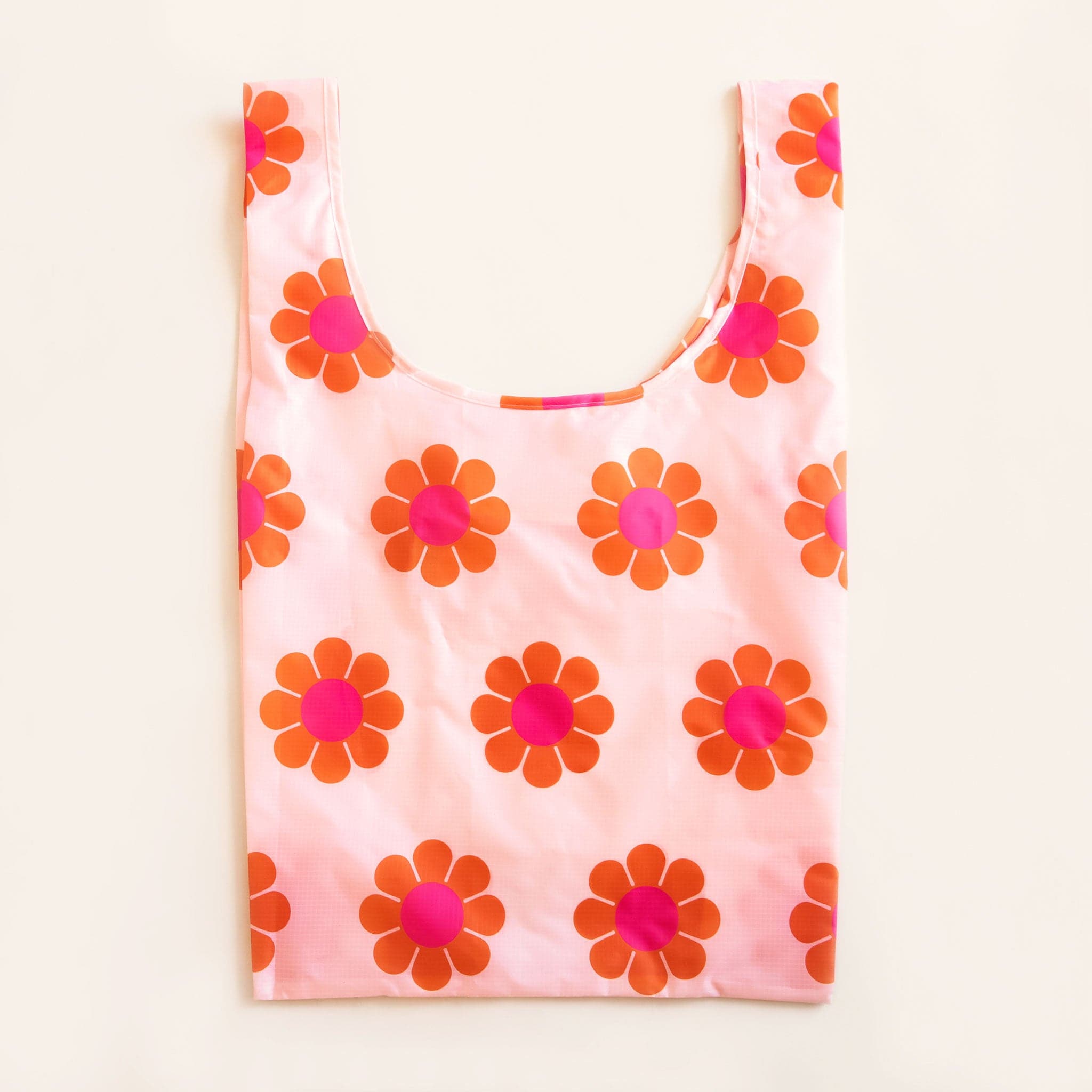 Peach reusable bag covered in a print of simple flowers with red-orange petals and magenta centers. The bag is positioned flat on a table and has a &#39;U&#39; shape between two handles.