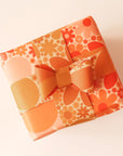Wrapped present covered in retro-inspired yellow and orange floral wrapping paper. Thin strips of wrapping paper with smaller similar print stretch around the present and fasten into a bow.