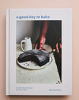 A white book cover with a photograph of a chocolate cake being cut along with the title that says, "a good day to bake" in light blue font.