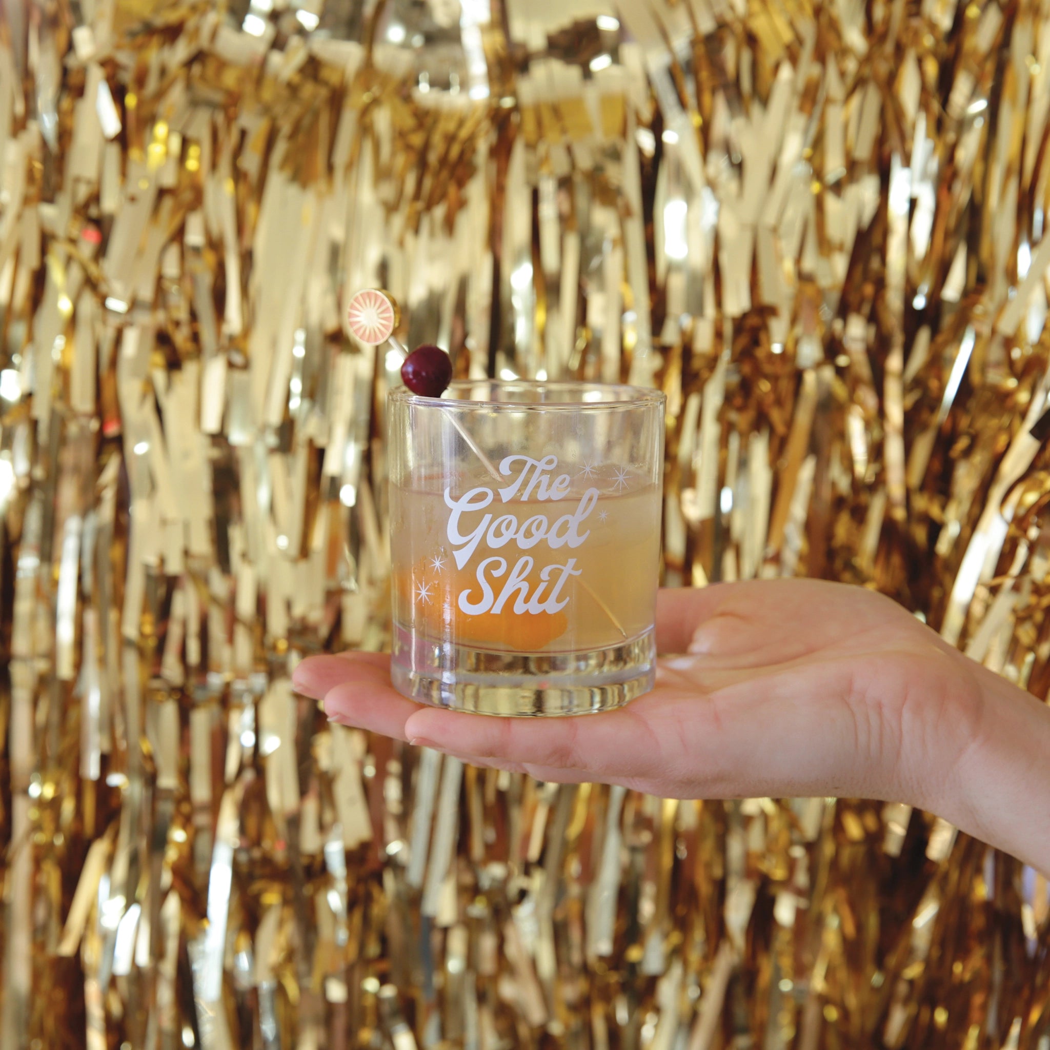 Against a gold tinsel backdrop is a photograph of a short glass tumbler with a thick bottom and &quot;The Good Shit&quot; printed across the center in white groovy cursive text.
