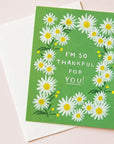 A photograph of a green card and a white envelope behind it. The card has a white daisy border around white text in the center that reads, "I'm So Thankful For You!".