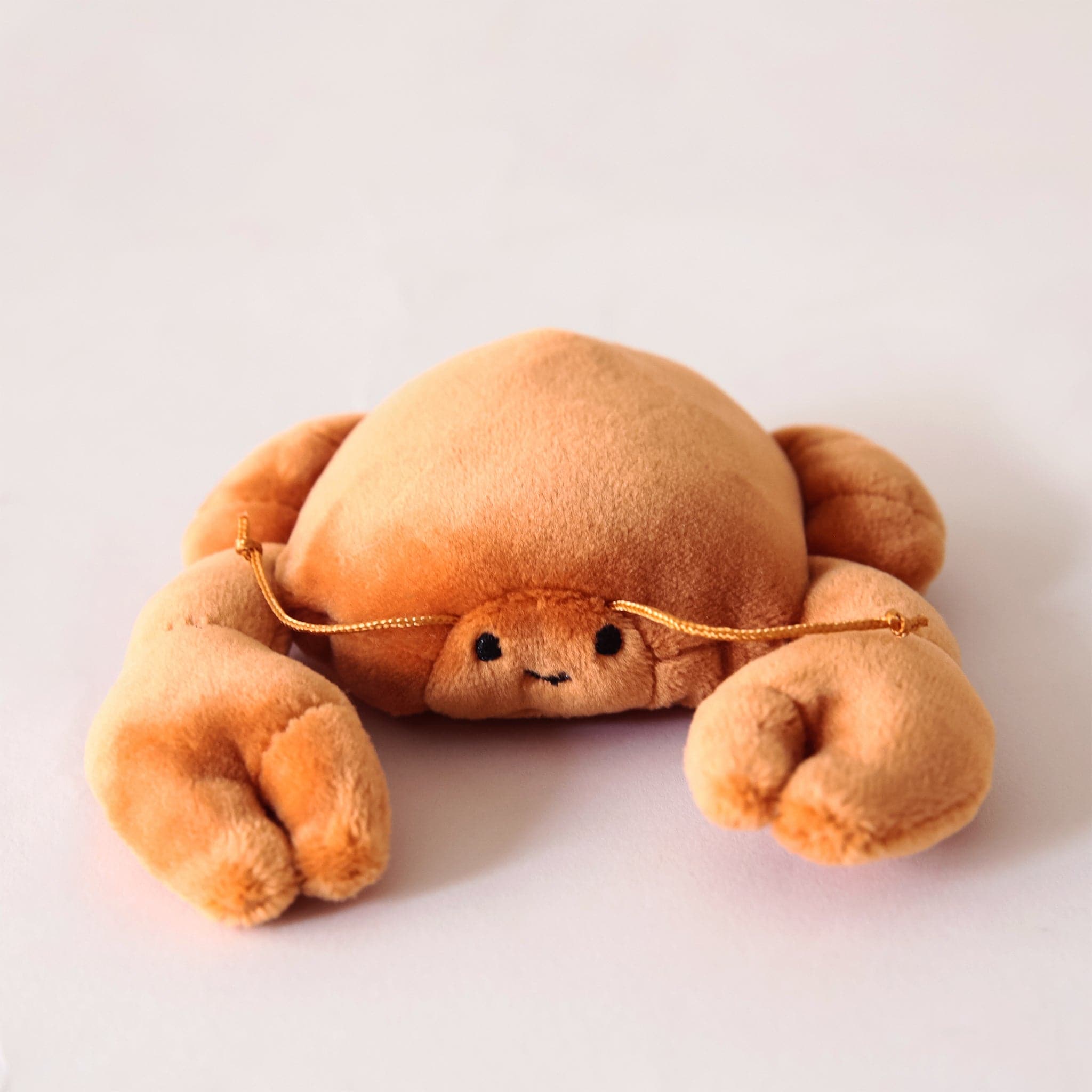 In front of a cream background is a stuffed orange crab laying on its stomach. It has two large claws in the front. On its face are two black eyes and a little black smile. He has two orange antennas above his eyes. 